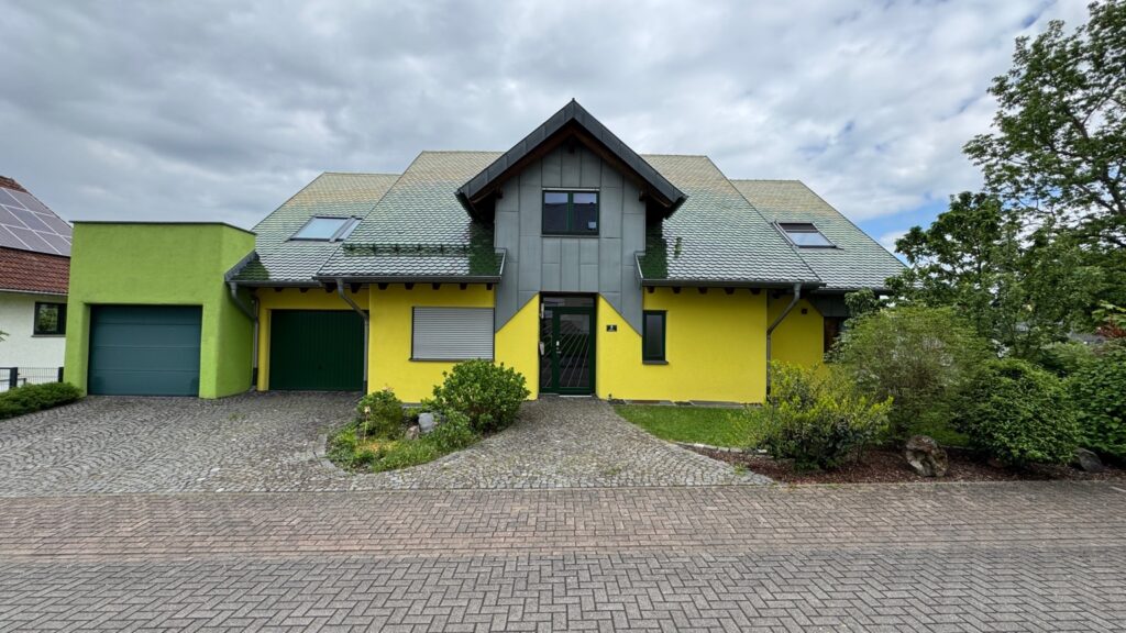 Vorderansicht der Ferienwohnung Greenheart: Modernes Einfamilienhaus mit gelber und grüner Fassade, zwei Garagen, gepflegtem Vorgarten und gepflasterter Einfahrt, unter bewölktem Himmel.